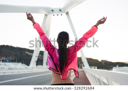 Back view of happy female with slender figure standing on a city bridge with raised hands while enjoying beautiful day after training, young sporty woman dressed in tracksuit relaxing after workout