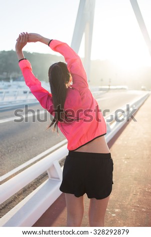 Back view of a young female jogger dressed in bright sportswear doing arms stretching exercise outdoors in summer day, athletic woman with perfect slim body working out while standing on running road