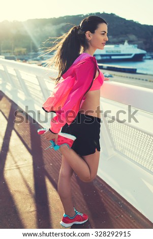 Full length portrait of a young sporty female dressed in colorful bright tracksuit doing stretching exercises for legs, beautiful fit woman engage physical activity outdoors in windy summer evening