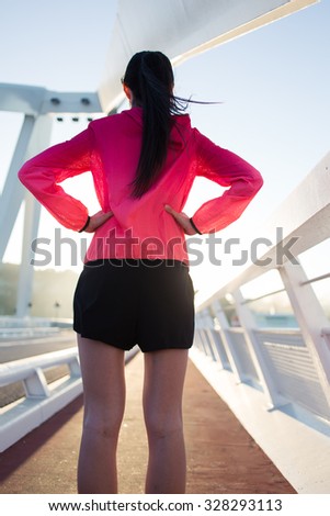Back view of young fit woman dressed in sportswear relaxing after workout outdoors in summer evening, sporty girl having rest after physical exercise outdoors while standing and admiring sunset