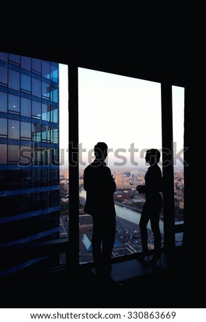 Silhouette of two confident colleagues having conversation while standing in corridor big company near window in evening time, young skilled workers discuss ideas after business meeting with partners