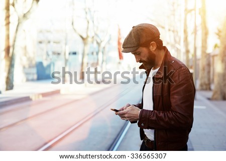 Half length portrait of bearded hipster man dressed in stylish clothes chatting on cell telephone while standing in the street, glamorous male with cool style use smart phone during strolling outdoors