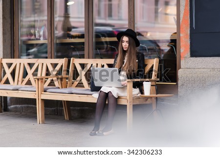 Portrait of glamorous hipster girl sitting with portable laptop computer on a cozy bench near coffee shop, young funky woman with nice look connects to the Internet via net-book though cafe wireless