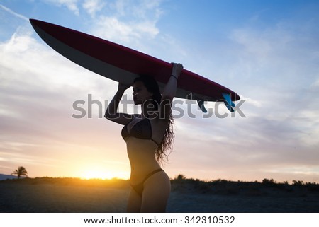 Silhouette of female model with curvy figure carrying surfboard above her head while walk on the beach at colorful sunset, young woman enjoying warm summer day on seashore while preparing for surf