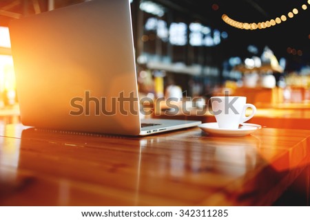 Open laptop computer with space for your brand lying on a wooden table in modern cafe bar interior, portable net-book and cup of hot drink, electronic distance work via internet during coffee break