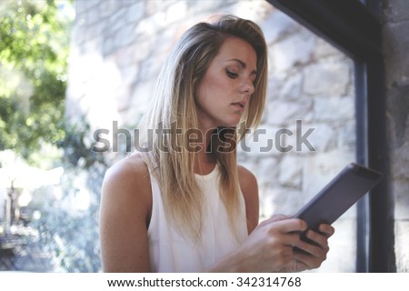 Portrait of a young beautiful blonde women reading electronic book on her digital tablet while standing near big window in office space, charming Sweden female using touch pad during work break