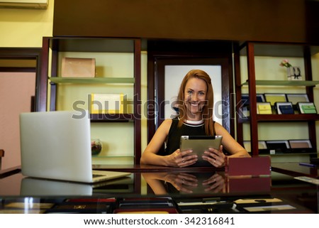 Portrait of a charming happy business woman holding digital tablet while standing near open laptop computer at cashier counter, young female seller with beautiful smile using touch pad and net-book