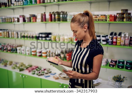 Young beautiful female consultant checks the quality of spa goods verifying information on digital tablet, woman owner using touch pad for work while standing in cosmetics store or pharmacy interior