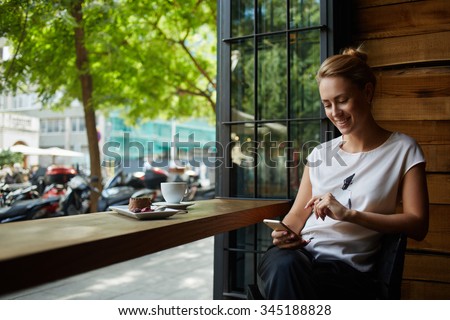 Charming woman with beautiful smile reading good news on mobile phone during rest in coffee shop, happy Caucasian female watching her photo on cell telephone while relaxing in cafe during free time