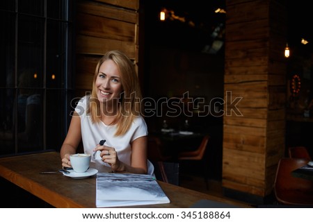 Attractive happy hipster girl with good mood posing while sitting alone in modern coffee shop interior, cheerful Caucasian woman with beautiful smile enjoying her recreation time in cozy cafe bar