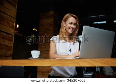 Pretty female student with cute smile keyboarding something on net-book while relaxing after lectures in University, beautiful happy woman working on laptop computer during coffee break in cafe bar