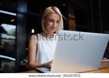 Beautiful young blonde female student using portable laptop computer while work at the coursework, woman sitting at the table with open net-book in coffee shop interior during morning breakfast
