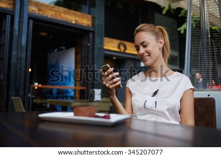 Young happy female reading good news on her mobile phone while sitting in modern coffee shop interior, gorgeous hipster girl with beautiful smile watching funny video on cell telephone during lunch