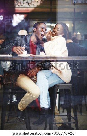 View through cafe window of a young happy dark skinned man and woman having fun while sitting together in bar, cheerful smiling black couple enjoying recreation time while having lunch in coffee shop