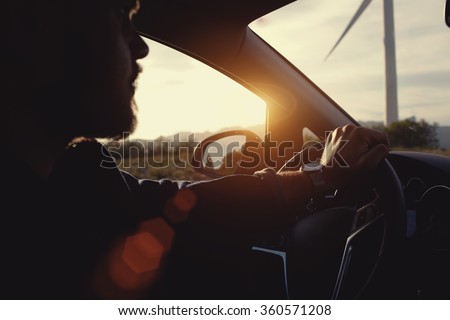 Young bearded hipster guy sitting behind the wheel of rented car during his anticipated travel on summer vacations, man riding in automobile at the countryside with beautiful evening sky and sun rays