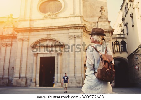 Back view of a young female wanderer out sightseeing in a foreign city during weekend overseas, trendy woman traveler with a rucksack on her back walking on unfamiliar street during summer adventure