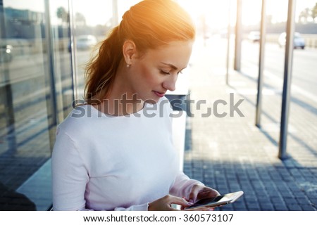 Young beautiful woman chatting with her friend via mobile phone while sitting on a bus stop in the city,charming hipster girl writing text message on her cell telephone while standing in urban setting