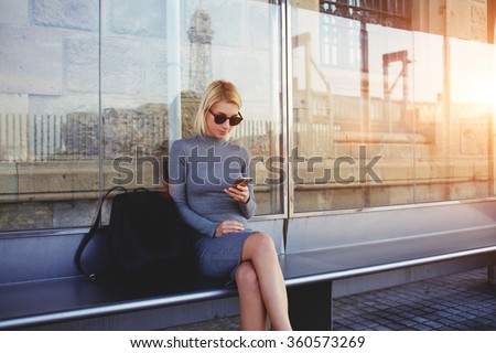 Stylish hipster girl chatting in network via smart phone with her friends while sitting on bus stop in summer day,fashionable woman tourist searching information about bus traffic using cell telephone