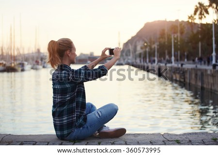 Woman posing while photographing herself on mobile phone for chat with friends in summer weekend, hipster girl enjoying her travel while making photo of beautiful landscape with cell telephone camera
