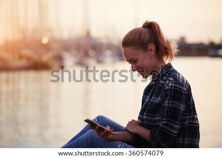 Young cheerful hipster girl sitting on a pier near sea port and read pleasant text message from her best friend, happy woman with beautiful smile chatting on cell telephone while relaxing outdoors
