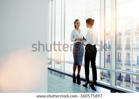 Businesswomen dressed in formal wear talking about work while meeting in hallway company, two female intelligent managers discussed plan the future conference while standing near big office window