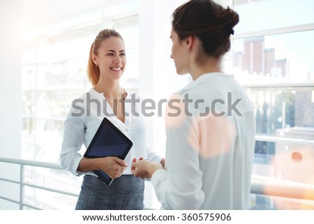 Smiling businesswoman holding digital tablet and talking with partner while standing in modern office interior, team of professional employees discussing ideas of project after working on touch pad
