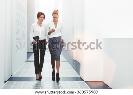 Confident women reading information about finance news while walking in company hallway during work break, successful businesswoman writing text message her client while goes with secretary to office