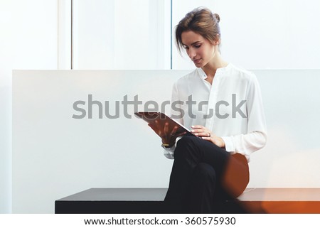 Young confident woman entrepreneur chatting on digital tablet with client while sitting on bench in office interior, intelligent female lawyer in formal wear reading electronic book during work break