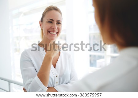 Two female entrepreneurs dressed in formal wear laughing at funny story after hard work day, young woman financier with beautiful smile listening her colleague while standing in modern office interior