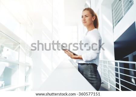 Young woman lawyer dreaming about something while standing with portable touch pad in modern office interior, female skilled managers thinking about new business ideas while work on digital tablet