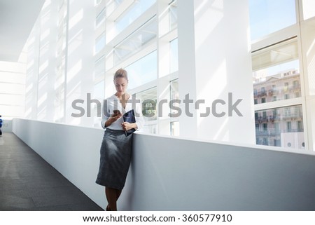 Elegant woman using mobile phone after work on digital tablet while standing in office interior,female manager read text message on cell telephone while waiting for her colleague after finish work day
