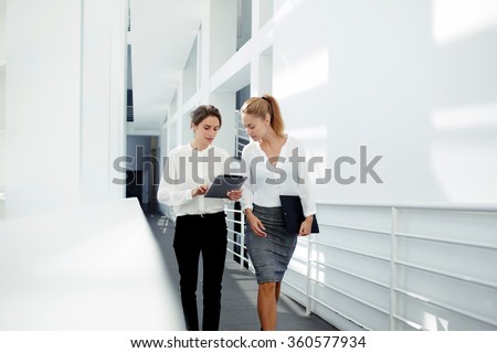Two female managing directors discussing ideas of project on digital tablet while walking down in office hall, confident women entrepreneurs working on touch pad while going to the conference room