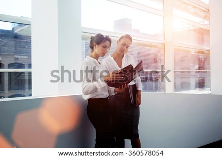 Successful women financiers discussing ideas of own project while standing in modern office interior, young female manager consulting with colleague about presentation while holding folder documents