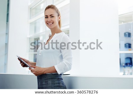 Smiling female managing director holding digital tablet while standing in modern office interior during work break, cheerful businesswoman holding touch pad while preparing for the important meeting
