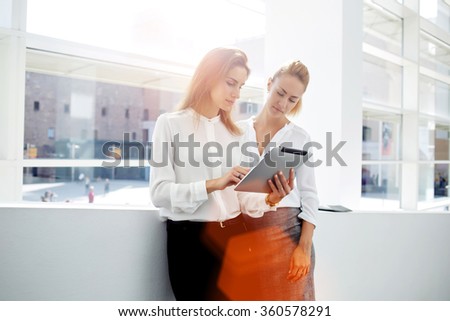 Two successful businesswomen checking list of affairs on digital tablet while standing in office interior, two female partners met in the hallway and stopped for consult new project on touch pad