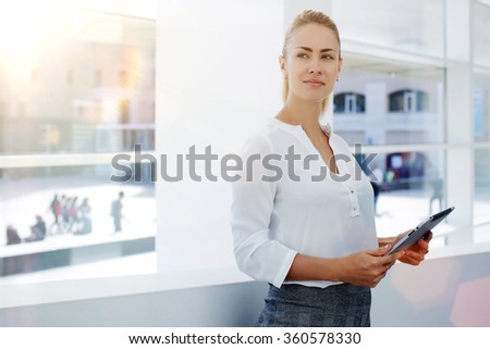 Young businesswoman holding touch pad and thinking about forthcoming meeting with important partners, female intelligent lawyer standing with digital tablet in modern office interior during work break