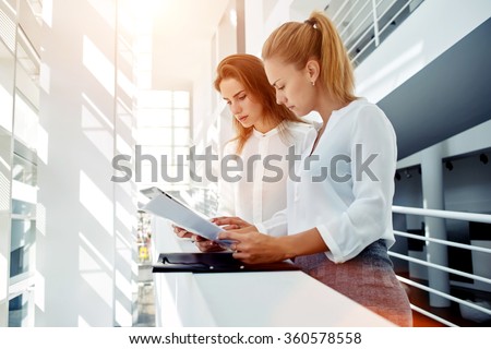 Couple of a businesswomen reading paper documents and using touch pad for prepare to meeting with partners, two female CEO check information on digital tablet while they standing in office interior