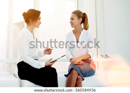 Young woman entrepreneur hold touch pad while telling something to her partner during business meeting, two female confident proud CEO discussing new project while sitting in modern office interior