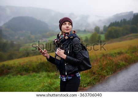Trendy woman catches signal on her mobile phone while searching interesting places in mountains, beautiful female tourist with rucksack waiting for a text message on cell telephone during walking tour