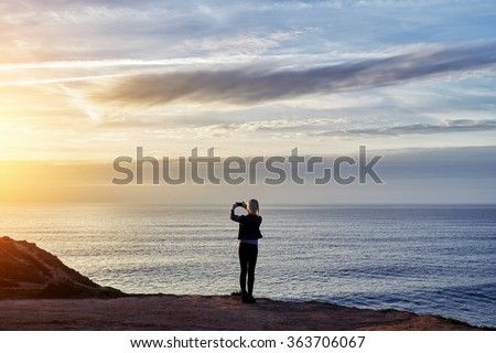 Back view of woman photographing amazing sunset with mobile phone while standing against copy space sea and sky background, hipster girl shoots video of a calm ocean and sunset on mobile phone