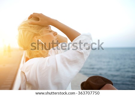 Half length portrait of young charming woman in headphones relaxing while sitting near ocean in summer evening, attractive carefree female listening favorite music during recreation time in weekend