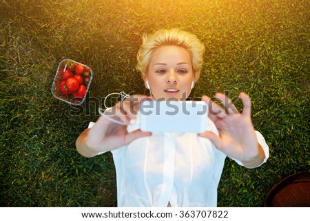 Top view of pretty young woman making self portrait on mobile phone while resting on grass plot in summer day, attractive female student taking picture with cell telephone camera while lying on a lawn