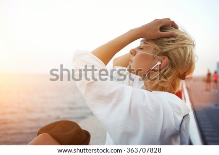 Beautiful blonde hair woman listening to music or radio in headphones while enjoying warm summer evening, pretty hipster girl with closed eyes resting on pier near sea during her long-awaited vacation