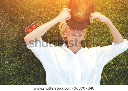Cheerful young woman closes own eyes from the sunlight using stylish hat while enjoying summer vacation, hipster girl with beautiful smile hiding face under headgear while lying on the grass in park