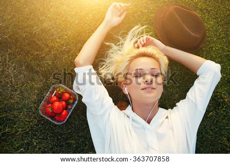 Top view of female student listening to music with headphones while lying on lawn after University lectures, young charming woman with closed eyes enjoying sunshine during recreation time in the park