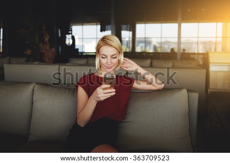 Gorgeous female reading news via network on mobile phone while waiting for her order in modern coffee shop, European woman searching information on cell telephone while sitting in restaurant interior