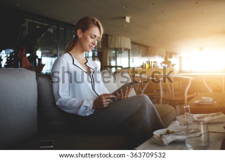 Young female entrepreneur reading electronic book on digital tablet during lunch in modern restaurant interior, elegant businesswoman chatting on touch pad while sitting in comfortable coffee shop