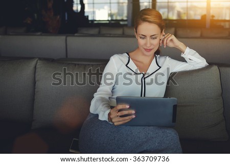 Young beautiful woman reading electronic book via internet on digital tablet while relaxing in coffee shop after work day, modern female watching fashion news on touch pad while waiting order in cafe