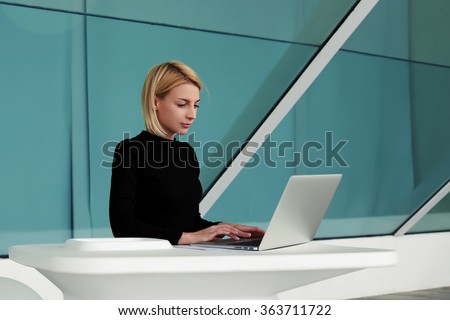 Concentrated woman developer performs software engineering on net-book while sitting in office interior, young businesswoman searching information in internet via laptop computer during work break