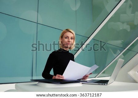 Young woman skilled manager holding paper documents while sitting an the table with open net-book, female secretary with resume of new employees in hands looking at camera in modern office interior